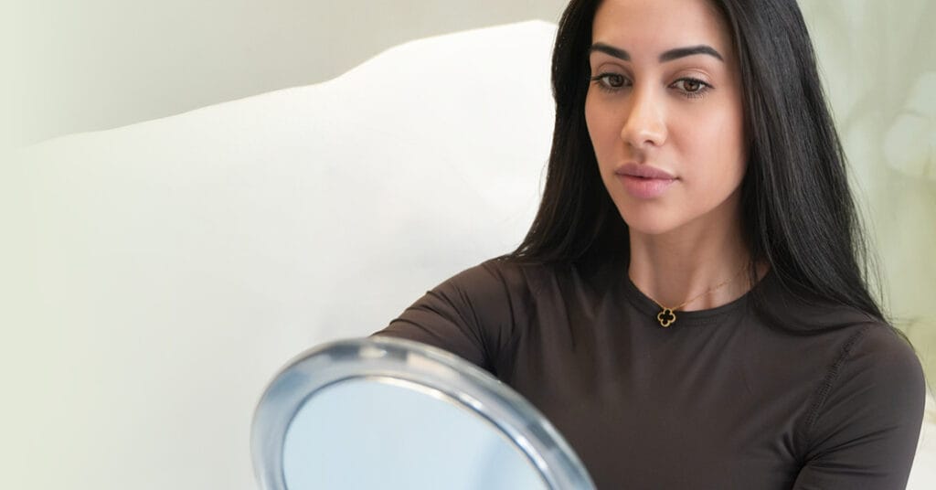 Glückliche Beauty2Go Patientin betrachtet Ergebnisse im Spiegel, nach minimalinvasiver Schönheitsbehandlung für frische, leuchtende Haut im Herbst